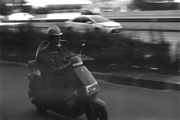 Ilford HP5 Plus 400 (FF)

English Alt Text

A black‑and‑white photo showing a person riding a scooter or small motorcycle in the foreground. The rider wears a helmet and appears slightly blurred, suggesting motion. Behind them, a car travels along a road, also somewhat blurred. The background includes trees and roadside elements, but details are indistinct due to the motion effect. The overall scene conveys everyday traffic movement.

中文替代文字

一张黑白照片，前景是一位戴头盔的人骑着电动车或摩托车，画面略微模糊，显示出运动感。后方有一辆汽车在道路上…
