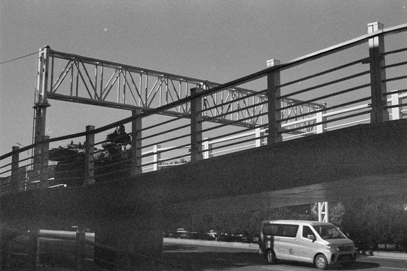 Ilford HP5 Plus 400 (FF)

English Alt Text

A black‑and‑white photo of a pedestrian bridge with metal railings and overhead beams. A person is pushing a cart up the ramp of the bridge. Below the bridge, a white van drives along a road. The van has text and a logo on its side, though not fully readable. The sky is clear, and shadows indicate sunlight. The scene highlights the structure of the bridge and the contrast between pedestrian space and road traffic.

中文替代文字

一张黑白照片，画面为带金属栏杆和上方横梁的人行天桥。桥上…