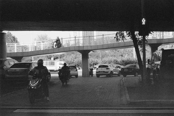 Ilford HP5 Plus 400 (FF)

English Alt Text

A black‑and‑white photo taken under an overpass. A pedestrian bridge crosses above, with a cyclist riding across it. Below, several parked cars line the side, and two people ride scooters or motorcycles along a paved path. A pedestrian signal on the right shows a lit walking figure. Trees and buildings appear in the background. The scene captures different forms of urban transportation in the same space.

中文替代文字

一张黑白照片，拍摄于高架桥下。上方的人行桥上有一名骑自行车的人经过。下方道路…