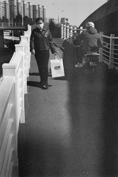 Ilford HP5 Plus 400 (FF)

English Alt Text

A black‑and‑white photo of a pedestrian walkway with white railings. On the left, a person wearing a face mask walks while carrying a shopping bag printed with Chinese characters and the words “TOP SPORTS.” On the right, a person rides a motorbike, wearing a helmet and jacket. Tall residential buildings and an elevated roadway appear in the background. The scene shows everyday movement in an urban environment.

中文替代文字

一张黑白照片，画面为带白色栏杆的人行步道。左侧一名戴口罩的人手提…