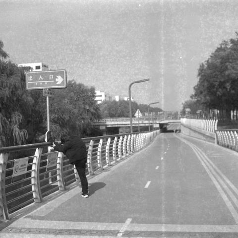 Ilford HP5 Plus 400 (6x6)

English alt text

A black‑and‑white image of a person stretching by placing one leg on the railing of a pedestrian path beside a road. The person wears dark clothing. The path has lane markings and is bordered by metal railings. Trees line both sides, and buildings appear in the background. A sign with Chinese characters is mounted on a pole nearby.

中文替代文字

一张黑白照片，一名身穿深色衣物的人将一条腿放在栏杆上做伸展动作。该步道位于道路旁，有车道标记并由金属栏杆围护。两侧种有树木，背景中可见建筑物。附近的路牌上有中文文字。