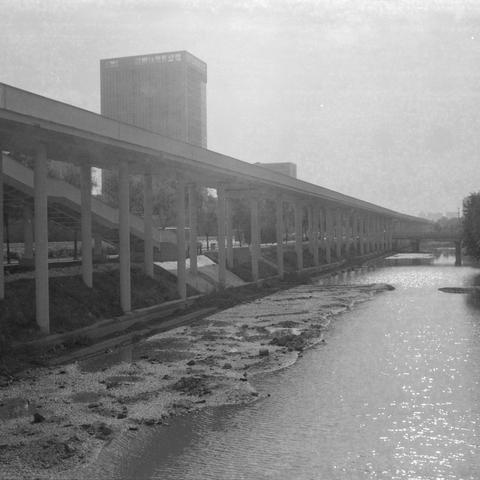 Ilford HP5 Plus 400 (6x6)

English alt text

A black‑and‑white image showing an elevated pedestrian walkway or rail structure supported by concrete pillars running alongside a shallow waterway. The water level is low, exposing mud and rocks along the banks. In the background stands a tall rectangular building with a grid of windows. Trees grow near the base of the structure. The scene captures urban infrastructure and a partially dry riverbed.

中文替代文字

一张黑白照片，画面是一条由混凝土柱子支撑的高架步道或轨道，沿着一条水位较低的河道延伸…