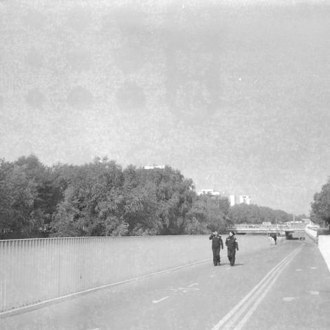 Ilford HP5 Plus 400 (6x6)
English alt text

A black‑and‑white photograph of two people in uniform walking side by side on a paved path marked with double yellow lines and a bicycle symbol. A metal railing runs along the left side, and trees line both sides of the path. Buildings are visible in the distance. The setting resembles an urban park or riverside walkway.

中文替代文字

一张黑白照片，两名穿制服的人并肩走在一条铺装道路上。道路上有双黄线和自行车标志。左侧有金属栏杆，两侧种有树木，远处可见建筑物。整体环境像是城市公园或河边步道。