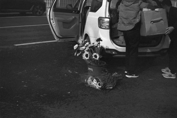 Ilford FP4 Plus 125 (FF)

English alt text
A black‑and‑white photo showing the back of a vehicle with its rear hatch and a side door open. Two people are lifting or lowering a large cardboard box into the back of the car. On the ground nearby, there is a bucket filled with sunflowers and a wrapped bouquet. Another vehicle is parked in the background. The scene appears to take place on a paved outdoor area.
中文替代文字
一张黑白照片，画面中一辆车的后备箱和侧门敞开着。两个人正把一个大纸箱搬进车内或从车里搬出。地面上放着一桶向日葵和一束用塑料包裹的花。背景中还能看到另一辆车，场景位于…