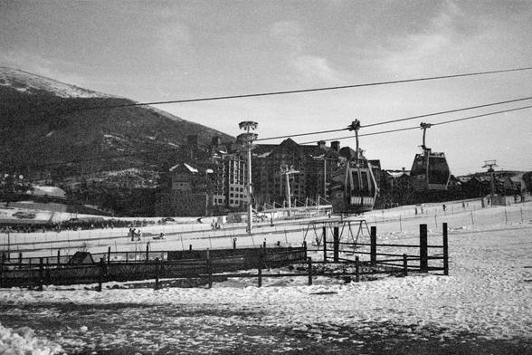 Ilford FP4 Plus 125 (FF)

English alt text
A black‑and‑white photo of a snowy ski resort. Several gondola cabins hang from overhead cables, moving above open ski slopes. A few people are visible on the snow, either skiing or walking. In the background, large multi‑story lodge buildings sit at the base of a mountain. A fenced area occupies the foreground. The scene appears calm and functional, showing typical winter recreation infrastructure.
中文替代文字
一张黑白照片，画面为积雪覆盖的滑雪场。多辆缆车吊舱悬挂在空中的钢索上，越过滑雪坡道。几个人在…