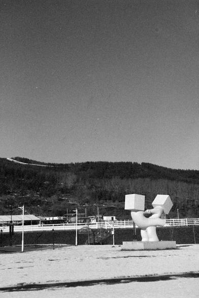 Ilford FP4 Plus 125 (FF)

English alt text
A black‑and‑white photo of an outdoor area featuring a modern abstract sculpture. The sculpture consists of two large cube‑like blocks connected by intertwined, rounded forms, all mounted on a rectangular base. The ground appears lightly covered with snow or pale sand. A simple fence and several poles surround the sculpture. In the background, there is a hillside covered with trees and a few small buildings. The overall scene looks quiet and open.
中文替代…