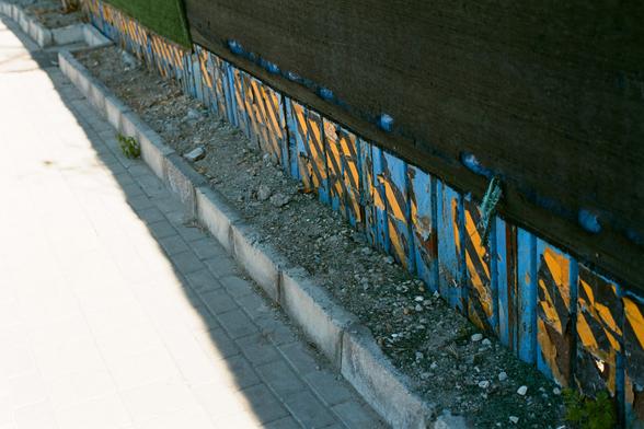 Lucky  C200 (FF)
English:

A close-up of a weathered, blue and yellow wooden fence running alongside a paved sidewalk. The fence is partially broken and peeling, with some slats missing or damaged. The sidewalk is made of concrete pavers, and there is a narrow strip of dirt and small rocks between the fence and the pavement. The scene is lit by natural sunlight, casting shadows on the ground.
Chinese:

一个靠近人行道的蓝黄色木栅栏特写。木栅栏部分破损、剥落，有些木条缺失或损坏。人行道由混凝土铺成，栅栏和路面之间有一条狭窄的土带和小石子。阳光照射下，地面上投射着影子。