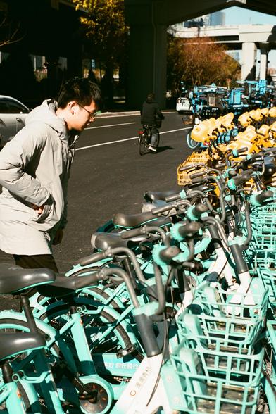 Lucky  C200 (FF)
English:

A person wearing a light gray jacket and glasses is standing next to a row of parked shared bicycles. The bicycles are turquoise and yellow, and the person seems to be looking at something on the ground. The background shows another cyclist and a street with vehicles.
Mandarin Chinese:

一位穿着浅灰色夹克、戴眼镜的人站在一排共享单车旁。单车为青绿色和黄色，该人似乎在看地上的某物。背景中有一位骑行者和有车辆行驶的街道。