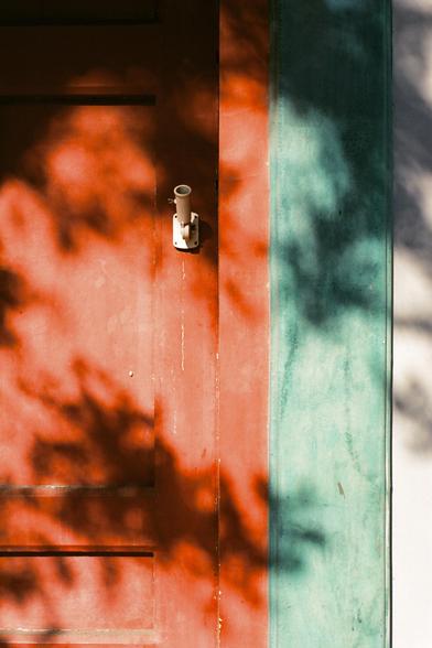 Lucky  C200 (FF)

English:
A close-up of a red door with a white cylindrical object. The door is partially illuminated by sunlight, casting a warm orange glow and creating blurred shadows of tree leaves on its surface.

中文:
一扇红色门的特写，门上有一个白色圆珠形物体。阳光部分照亮了门，投射出温暖的橙色光芒，并在门面上形成树叶模糊的影子。