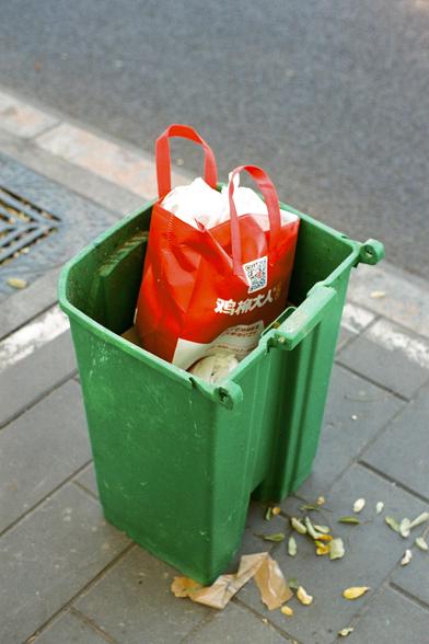 Lucky  C200 (FF)

English:
A green trash bin on a sidewalk, containing a red plastic bag filled with items. The bag has white text and a QR code. Some leaves and debris are scattered around the base of the bin.

中文:
人行道上有一个绿色垃圾桶，里面有一个装满物品的红色塑料袋，塑料袋上有白色文字和二维码。垃圾桶底部周围散落着一些树叶和碎片。