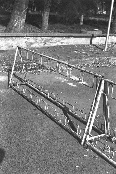 Ilford FP4 Plus 125 (FF)

English: A black-and-white photograph of an empty metal bike rack lying on its side on a paved surface. The bike rack is triangular in shape with multiple horizontal bars for securing bicycles. In the background, there are trees, a bench, and a low wall.

中文: 这是一张黑白照片，照片中一个空的金属自行车架倒在地上，位于铺砌的路面上。自行车架呈三角形，有多个水平横杆用于固定自行车。背景中有树木、长椅和一堵矮墙。