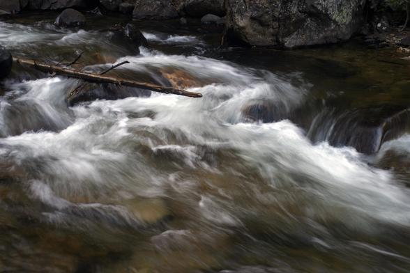 A cataract along Boulder Brook in Rocky Mountain National Park. The brook descends from near the top of Storm Peak down to Storm Pass, a spot along Bear Lake Road uphill from Sprague Lake. A trail follows the brook and this spot caught my eye. The brook is about 15’ across and a lot of water is rushing over some rocks throwing up white water the looks silky with a long exposure. A thin log extends from the left edge as though suspended over the water. September 2003