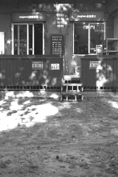 Ilford FP4 Plus 125 (FF)

English:
A black-and-white photograph of a storefront with signs in Chinese. The signs advertise services such as printing, copying, and photo development. A small stool or step ladder casts a shadow on the ground in front of the store.

Mandarin Chinese:
一张黑白照片，展示了一个中文招牌的店铺门面。招牌上提供打印、复印和照片冲印等服务。店铺前地面上有一个小凳子或脚凳的阴影。