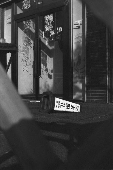 Ilford FP4 Plus 125 (FF)

English: A black-and-white photograph showing a storefront with glass doors. The glass is sticked with torn posters and may be partially broken. In front of the store, there is a knocked-over traffic cone with text written in Chinese characters.
中文: 这是一张黑白照片，展示了一家有玻璃门的店铺。玻璃门上贴满被撕毁的告示，可能部分破碎。店铺前有一个被撞倒的交通锥，上面写着中文字。