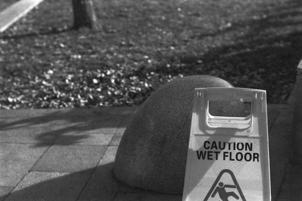 Ilford FP4 Plus 125 (FF)

English:
A black-and-white close-up of a "Caution Wet Floor" sign placed on a paved walkway. The sign is positioned next to a large rock, with a landscaped area and trees in the background.

中文（普通话）：
一张黑白照片，特写镜头拍摄了一个“小心地滑”的警示牌，放置在铺砌的人行道上。警示牌旁边有一块大石头，背景是景观区和树木。