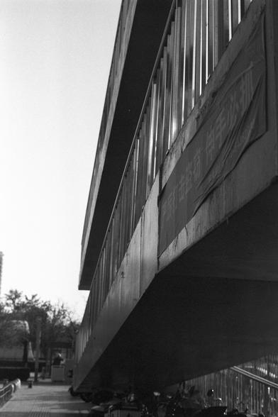 Ilford FP4 Plus 125 (FF)

Image 1:
This is a black-and-white photograph of a pedestrian over walk. The walk has a unique architectural design with sharp, angular lines and metal railings. The structure is elevated, supported by pillars, and there is a sidewalk underneath with bicycles parked along the side. The photo is taken from a low angle, emphasizing the geometric shapes and shadows of the building.

中文:
这是一张黑白照片，拍摄的是一座过街天桥。天桥具有独特的建筑设计，线条锐利，角度分明，并且有金属扶手。建筑物由柱子支撑，下方有一条人行道，沿路停放着自行车。照片从低角度拍摄，…