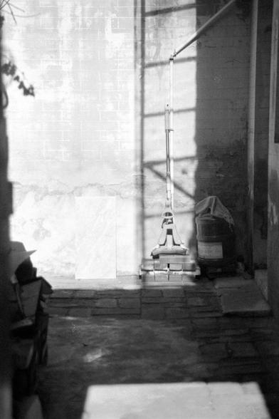 Ilford FP4 Plus 125 (FF)
English:

A black-and-white photograph of a semi-enclosed outdoor area. In the center, there is a tall floor polisher placed on a stack of bricks. To the right, there is a bucket with a cloth draped over it. The light from a window or opening casts shadows on the wall and floor, creating a contrast of light and shadow. The scene has an industrial or maintenance feel.
Mandarin Chinese:

这是一张黑白照片，拍摄的是一个半封闭的室外区域。中央有一个高大的地板打蜡机，放在一堆砖块上。右边有一个桶，上面搭着一块布。来自窗户或开口的光线在墙壁和地板上投射出影子，形…
