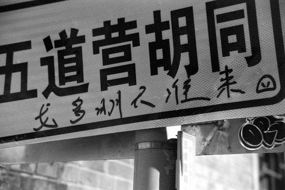 Ilford FP4 Plus 125 (FF)
English:

This is a black-and-white photograph of a signboard with Chinese characters. The main text on the signboard reads "五道营胡同," with additional handwritten characters below it read “戈多明天准来” translating to “Godot is definitely coming tomorrow”.
普通话:

这是一张黑白照片，照片中是一个写有汉字的标牌。标牌上的主要文字是“五道营胡同”，下方还有一些手写的汉字 “戈多明天准来”。