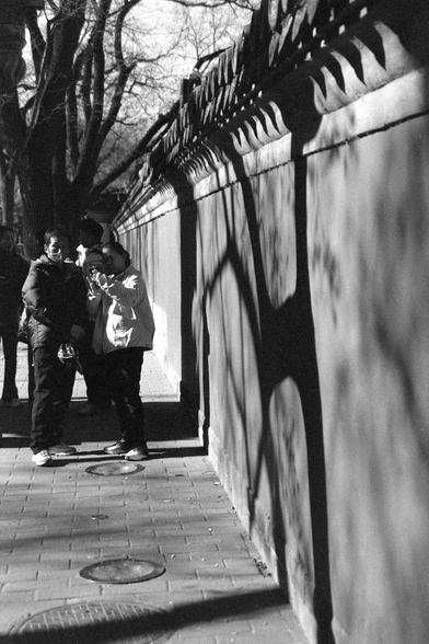 Ilford FP4 Plus 125 (FF)

##English
A black-and-white photograph of four people standing together near a decorative wall with pointed tops. The wall casts shadows on the ground, creating a pattern of light and dark. The people appear to be engaged in conversation or looking at something off-camera. The scene is set on a paved walkway, with trees in the background.

##中文
一张黑白照片，四个人站在一个带有尖顶装饰的墙壁旁。墙壁在地面上投射出阴影，形成明暗相间的图案。这三个人看起来在交谈或注视着镜头外的某物。场景设在铺砌的步道上，背景有树木。