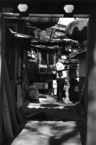 Ilford FP4 Plus 125 (FF)

##English
A black-and-white photograph of a person standing inside a dimly lit, cluttered workshop or storage area. The individual is wearing a jacket a helmet and glasses, standing near various objects and equipment. The ceiling is low and filled with wires and wooden stacks, and there are large lanterns hanging above. The scene is framed by a doorway, with light streaming in from outside.

##中文
一张黑白照片，一个人站在光线昏暗、杂物众多的车间或仓库内。这名个体穿着夹克，戴着安全帽和眼镜，站在各种物品和设备旁边。天花板低矮，布满电线和木板，…