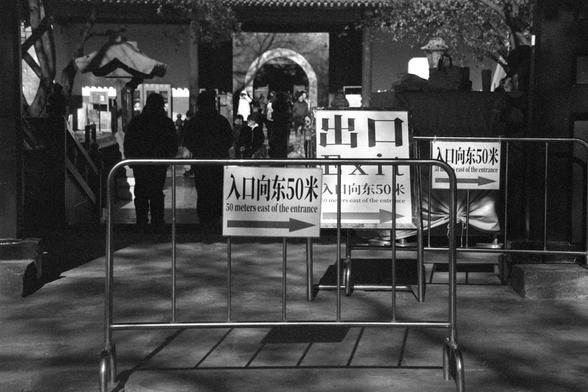 Ilford FP4 Plus 125 (FF)

##English
A black-and-white photograph showing a metal barricade with signs indicating "Entrance 50 meters east of the entrance" and "Exit." The signs are written in both Chinese and English. The barricade is set up to guide the flow of people, with several individuals visible in the background, walking toward an arched entrance.

##中文
一张黑白照片，展示了一个金属护栏，上面挂有标志牌，写着“入口向东50米”和“出口”，标志牌上有中英文。护栏用于引导人流，背景中有几个人朝拱形入口走去。