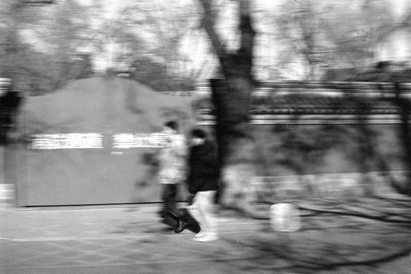 Ilford FP4 Plus 125 (FF)

English:
A black-and-white, motion-blurred photograph of two people walking on a sidewalk. The background includes trees and a wall with Chinese characters, though the text is blurred and unreadable due to the motion effect. The scene appears to be outdoors, possibly in an urban or park setting.

中文：
一张黑白且带有动感模糊的照片，拍摄的是两个人在人行道上行走。背景中有树木和一面写有中文字的墙，但由于动感模糊，文字无法辨认。场景看起来是在户外，可能是城市或公园环境