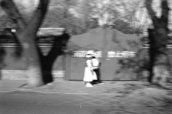 Ilford FP4 Plus 125 (FF)

English:
A black-and-white, motion-blurred photograph of two people walking outdoors. The background includes trees and a wall with blurred Chinese characters. The scene is similar to the previous image, with the motion blur creating a sense of movement.

中文：
一张黑白且带有动感模糊的照片，拍摄的是两个人在户外行走。背景中有树木和一面写有模糊中文字的墙。场景与上一张照片类似，动感模糊营造出运动感。