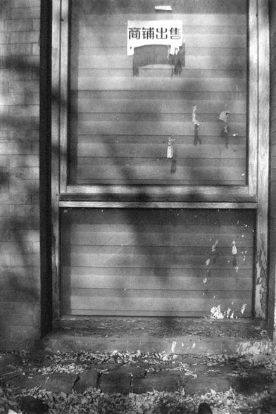 Ilford FP4 Plus 125 (FF)

##English
A black-and-white photograph of a wooden door with horizontal panels. The door is slightly weathered, with peeling paint and some dirt at the base. Above the door, there is a small rectangular window with a sign that reads "商铺出售" (Shop for Sale) in Chinese characters. The ground in front of the door is covered with fallen leaves and debris. The door is framed by brick walls on either side.

##中文
一张黑白照片，拍摄的是一扇带有横向木板的木门。门有些许风化，漆皮剥落，底部有灰尘和污渍。门上方有一个小长方形窗户，窗户上挂着一个…