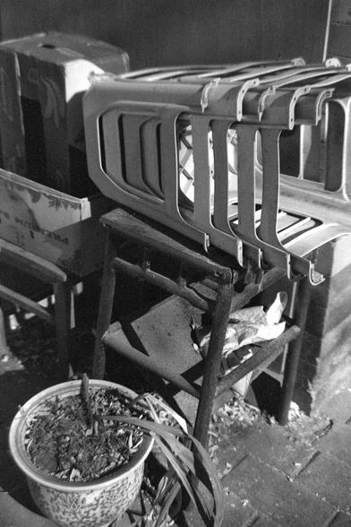 Ilford FP4 Plus 125 (FF)

##English
A black-and-white photograph showing a group of old, stacked plastic chairs with supports. The chairs are placed against a wall, and some are missing seats or have broken parts. In the foreground, there is a large potted plant with dry soil. The ground is paved with stone tiles, and there is dry leaves on the ground near the chairs.

##中文
一张黑白照片，照片中是一组堆放在一起的旧塑料椅子放在旧木椅上。部分椅子缺少座板或有损坏。前景处有一个大花盆，里面的土壤干燥。地面铺着石板，椅子旁边的地上有一些枯叶。
