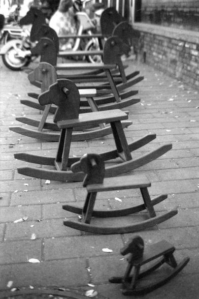 Ilford FP4 Plus 125 (FF)
English
A black and white photograph of several wooden rocking horses arranged in a row on a paved area. In the background, there are blurred figures of people and a motorcycle. A child is seen playing near one of the rocking horses.
中文
一张黑白照片，拍摄的是几个木制摇摆木马排成一排放在铺砌的区域。背景中有模糊的人影和一辆摩托车。一个孩子在其中一个摇摆木马附近玩耍。