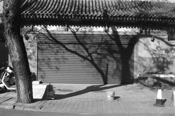 Ilford FP4 Plus 125 (FF)
English
A black and white photograph of a traditional building with a tiled roof and ornate eaves. The building has a closed metal shutter door, and shadows of tree branches are cast on the shutter and the ground. A tree is visible on the left side of the image. There are a few objects, including a bucket and a traffic cone, placed on the ground in front of the shutter.
中文
一张黑白照片，拍摄的是一座传统建筑，建筑有瓦片屋顶和精美的屋檐。建筑前有一个关闭的金属卷帘门，树枝的影子投射在卷帘门和地面上。照片左侧可见一棵树，卷帘门前的地面上放着几样物品，包括一个水桶和一个交…