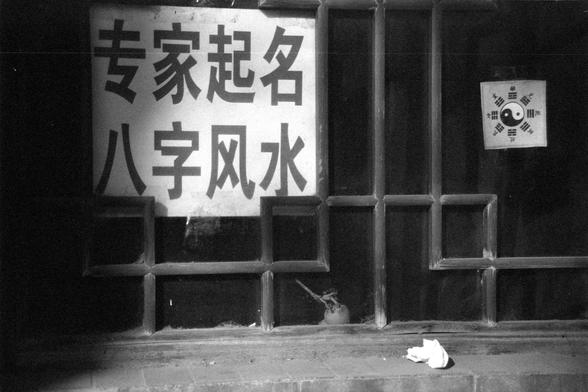 Ilford FP4 Plus 125 (FF)

English

A black-and-white photograph showing a wooden wondow with a large sign in Chinese characters. The sign reads "专家起名 八字风水" (Expert Naming, Eight Characters Feng Shui). To the right of the door, there is a small hanging sign with a Yin-Yang symbol and trigram patterns. On the window frame in the front of, there is a piece of cloth and a small object.

中文

一张黑白照片，展示一个木窗，窗上挂着一个大的中文招牌，写着“专家起名 八字风水”。门的右侧挂着一个小牌子，上面有阴阳符号和八卦图案。窗前有一块布和一个小物品。