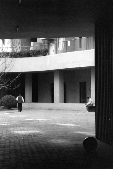 Ilford FP4 Plus 125 (FF)

English

A black-and-white photograph capturing a courtyard or open space between buildings. Two individuals are visible: one is walking away from the camera, and the other is sitting on a low wall or ledge. The buildings have large windows and balconies, and there are some trees and bushes in the background.

中文

一张黑白照片，拍摄的是建筑物之间的庭院或开放空间。照片中有两个人：一个人正背对镜头走开，另一个人坐在低矮的墙或台阶上。建筑物有大窗户和阳台，背景中有一些树木和灌木。