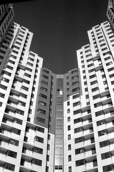 Ilford FP4 Plus 125 (FF)

English

A black-and-white photograph capturing a view looking up at two tall apartment buildings. The buildings have numerous windows and balconies, creating a symmetrical and repetitive pattern. The sky is visible between the buildings, and the perspective emphasizes the height and uniformity of the structures.

中文

一张黑白照片，拍摄的是仰视两栋高层公寓楼的视角。楼房有许多窗户和阳台，形成对称且重复的图案。两栋楼之间可见天空，视角强调了建筑的高度和统一性。