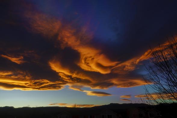 Clouds at sunset.
A dark silhouette of a horizon settles at the bottom of the image. The light blue sky immediately above it fades quickly to very dark blue at the top. Across the middle, billows of clouds that would be fluffy white at mid-day are instead washed in oranges and yellows and shadows. November 2017