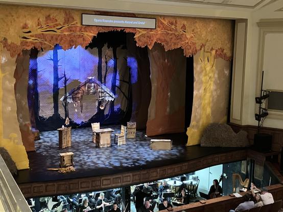 A stage set for the opera "Hansel and Gretel," featuring a backdrop of colorful trees and a rustic cabin. Several wooden props, including tables and chairs, are positioned on the stage. The orchestra is visible below in the pit.