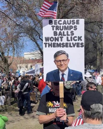 From No Kings March.
A person in the crowd is holding a sign with an image of a smiling (one might say, smirking) House Speaker Johnson, with text, “Because Trumps balls won’t lick themselves.”