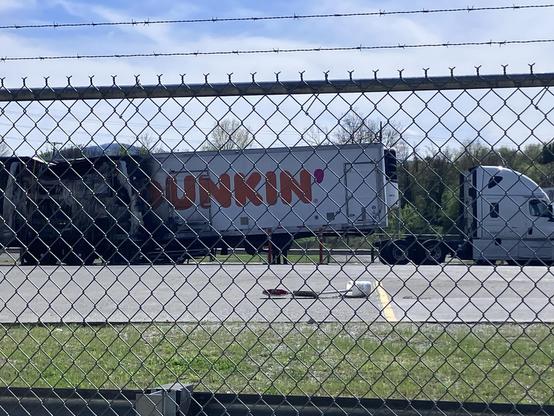 A burned-out truck trailer with the Dunkin' logo is parked next to an intact white truck inside a barbed wire fence. 