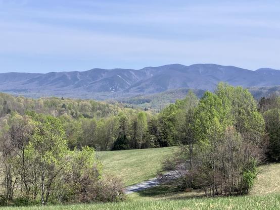 A scenic view of rolling hills and mountains in the background, with lush green trees and a grassy field in the foreground under a clear blue sky.