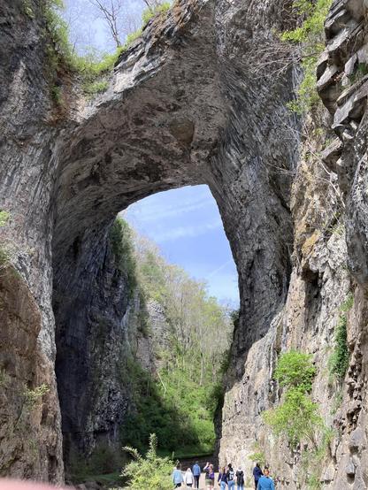 The Natural Bridge, a large rock arch towers above a lush, green valley, with people walking along a path beneath it. The scene features steep, rocky cliffs on either side, partially covered in vegetation, against a blue sky.