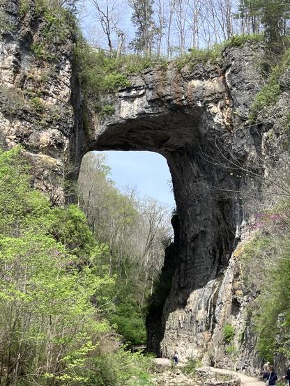 A natural rock arch stands tall amidst lush greenery and trees. A pathway runs through the area, with a few figures walking along it. The sky is clear, adding to the scenic beauty.