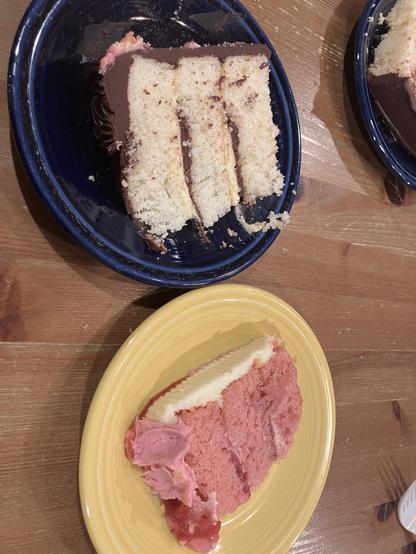 A blue plate contains a slice of Boston Cream cake with three layers. Next to it, a yellow plate holds a slice of strawberry cake topped with light icing and some strawberry filling.
