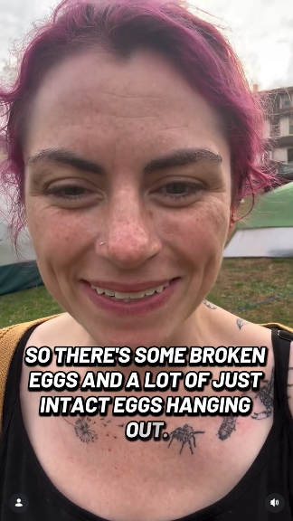 Still from the video: a middle aged white woman with tattoos and red-purple hair is grinning and talking about this nonsense in the video. The caption in this still reads "So there's some broken eggs and a lot of just intact eggs hanging out."