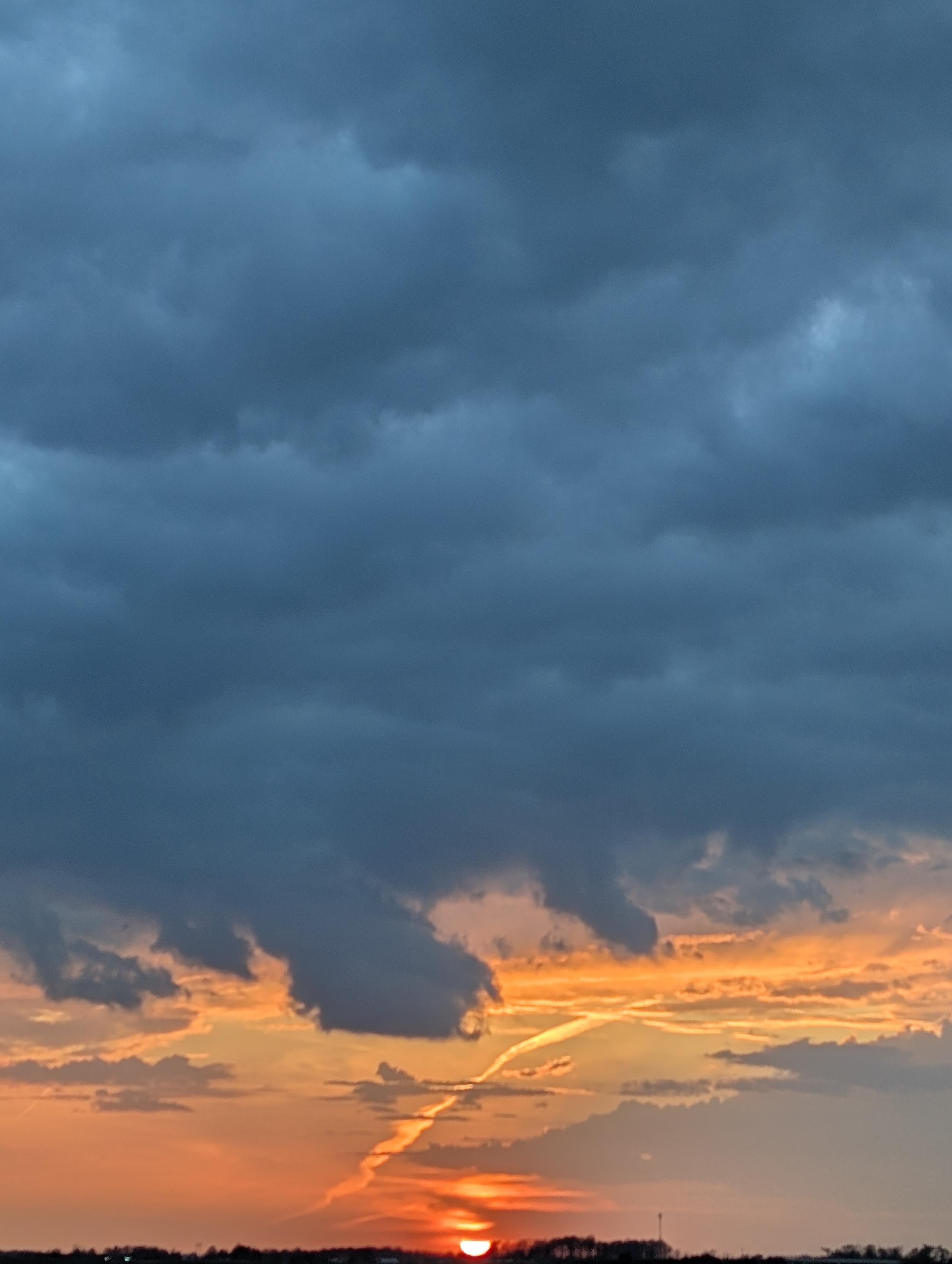 Sunset over the corn field. A sky full of blue clouds is interrupted by streaks of bright yellow and orange near the horizon. Patches of pale blue sneak through, providing contrast