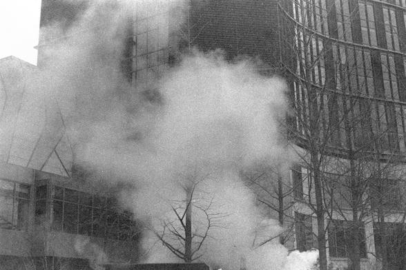 Ilford FP4 Plus 125 (FF)

English

A black-and-white photograph showing a building with thick smoke billowing out from several windows and the roof area. The smoke is dense and rises upward, partially obscuring the building's facade. The structure appears to be a multi-story building with scaffolding visible on the right side. Leafless trees are in the foreground, adding to the scene's stark atmosphere.

中文

一张黑白照片，展示了一栋建筑物，浓烟从多个窗户和屋顶区域滚滚冒出。烟雾浓密，向上升腾，部分遮挡了建筑物的外观。该建筑看起来是一栋多层建筑，右侧可见脚手架。前景中有光秃秃的树木…