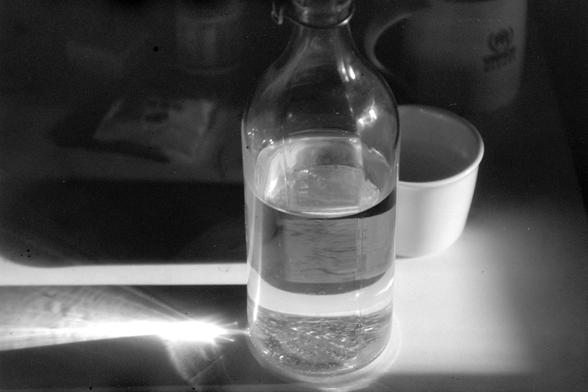 ERA 100 (FF)

English

A black-and-white photograph of a clear glass bottle, half-filled with liquid, placed on a surface. The bottle has a metal cap and a label wrapped around its middle. To the right of the bottle, there is a white bowl. The scene is well-lit, with light reflecting off the bottle and casting shadows.

中文

一张黑白照片，展示了一个透明玻璃瓶，瓶内装有半瓶液体，放置在一个平面上。瓶子带有金属盖，中间部分缠绕着标签。瓶子右侧有一个白色碗。场景光线充足，光线照射在瓶子上并投下阴影。