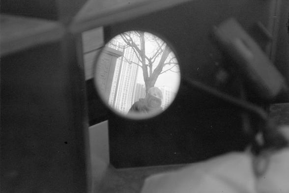 ERA 100 (FF)

English

A black-and-white photograph taken through a rearview mirror, showing a distorted view of a person with helmet, a tree and a building outside. The circular frame of the mirror is clearly visible, and the image appears slightly blurry due to the lens effect. The person is looking down. The tree is leafless, and the building in the background has vertical lines, possibly windows or architectural details.

中文

一张通过后视镜拍摄的黑白照片，展示了外面一个戴头盔的人，一棵树和一栋建筑物的扭曲视图。后视镜的圆形边框清晰可见，由于镜头效应，图像…