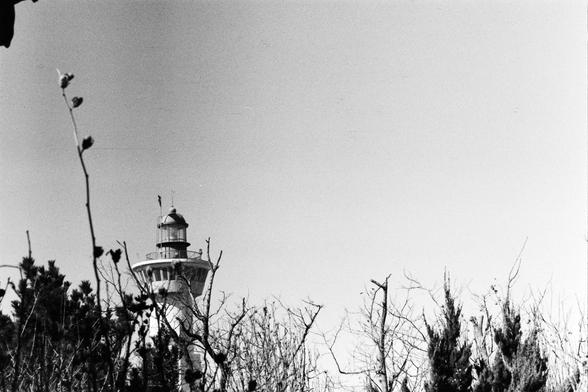 Lucky LUCKY SHD 400 (FF)

English

A black-and-white photograph showing the top of a lighthouse peeking above a line of trees. The lighthouse has a cylindrical base and a lantern room at the top. The sky is clear, and the trees in the foreground are bare, with a few sparse leaves.

中文

一张黑白照片，展示了一座灯塔的顶部从树林上方探出。灯塔底部为圆柱形，顶部是灯室。天空晴朗，前景中的树木枝叶稀疏，几乎没有叶子。
