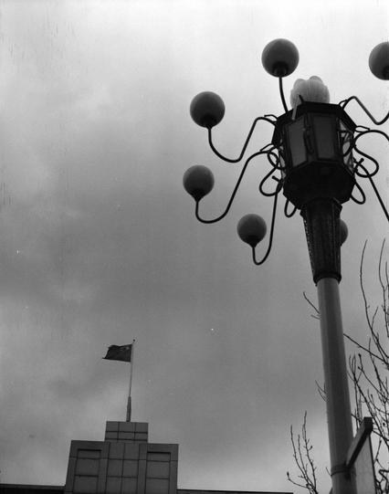 Lucky LUCKY SHD 400 (6x7)

English:

A black-and-white photograph of a street lamp with multiple round bulbs extending from its arms. In the background, a flag is flying atop a modern building with a flat roof and rectangular windows.

中文:

一张黑白照片，展示了一盏街灯，街灯有多个圆形灯泡从灯臂上延伸出来。背景中，一面旗帜在一座现代建筑的顶部飘扬，建筑物屋顶平坦，窗户为矩形。