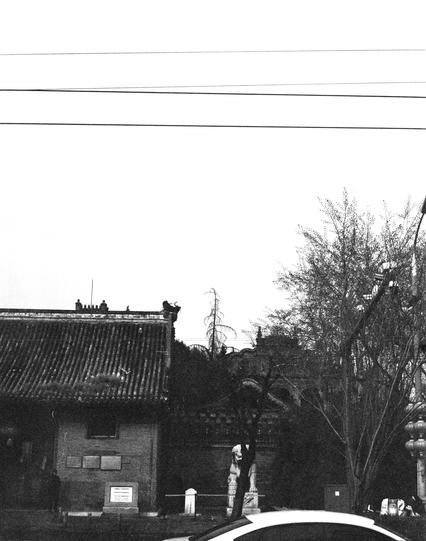 Lucky LUCKY SHD 400 (6x7)

English:

A black-and-white photograph showing a traditional Chinese building with a tiled roof and ornate eaves. The building is partially obscured by trees and power lines. A sign is visible on the wall, but the text is not legible. A car is partially visible in the foreground.

中文:

一张黑白照片，展示了一座传统中式建筑，建筑屋顶铺有瓦片，屋檐装饰华丽。建筑物部分被树木和电线遮挡。墙上有一个标志，但文字不清晰。前景中有一辆汽车部分可见。